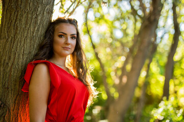 Poster - Pretty Caucasian woman leaning on a tree trunk with a red dress posing in the forest