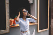 © BullRun - Stylish young ethnic lady in trendy sunglasses standing next to window of cafe and touching hair