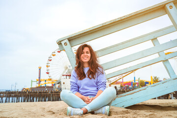 A young woman in a purple pullover sits on the sand at Santa Monica Beach. Los Angeles, USA 