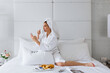 Young woman wearing bathrobe, drinking coffee and using mobile phone at bed in hotel room