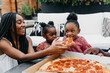 © Kristen Curette & Daemaine Hines/Stocksy - An African American family gathered around a table eating pizza.