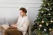 © Alina Hvostikova/Stocksy - Serious young guy using laptop sitting on sofa near decorated Christmas tree at home