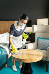 Poster - Young female in protective mask and uniform cleaning table in hotel room