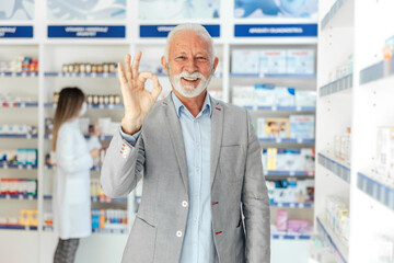 Wall Mural - Portrait of a male pharmacist showing a sign of approval with his hand, thumb up. A man with gray hair in a modern uniform and gesturing approval with one hand. Look at the camera, woman in background