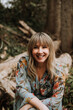 © Leah Flores/Stocksy - A Smiling Woman Sitting in a Woodsy Park Smiles and Looks at the Camera