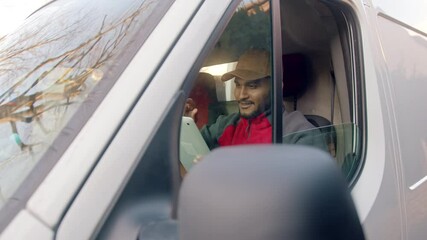 Wall Mural - Delivery guy wearing red uniform and a cap sitting in the front seat of the white van tipping his cap. Holding a tablet in his hands. Smiling and using tablet. Concept of Parcel Delivery. 