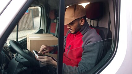 Wall Mural - Delivery boy wearing a cap sitting in the Driving seat of the van with a cardboard box placed on the other seat. Looking up address using a tablet. Concept of parcel delivery services. 