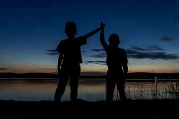 Silhouette of two children standing with their hands up on the shore of the lake at sunset. Family vacation by the lake, camping