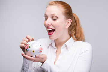 Wall Mural - Young happy red haired woman feed her piggy bank / porcelain bank with a coin