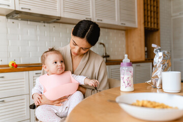 Young brunette mother smiling and feeding her baby in kitchen at home