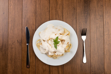 Tagliatelle pasta served with bechamel sauce and basil leaves. Pasta with white sauce. Top view gastronomic photography. Fork and knife beside white plate.