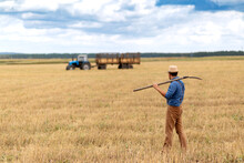 A Farmer Holds A Pitchfork And Looks At A Blue Tractor In A Field