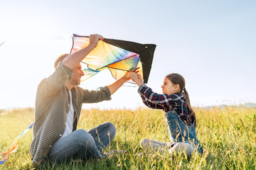 Smiling girl sitting on the grass with father helping her to prepare colorful rainbow kite toy for flying. Happy family childhood moments or outdoor time spending concept image.