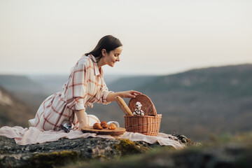 Wall Mural - Young woman sits on a rocky mountain at picnic with basket with croissants, wine and baguette.