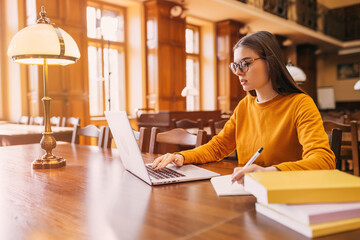. A young student sits on a chair at a table in the library, she studies and uses a laptop to take notes.