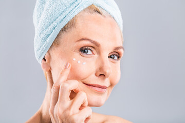 Senior smiling 50s middle aged mature older woman applying facial cream on face looking at camera isolated over white background. Anti age healthy dry skin care beauty therapy concept