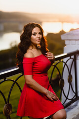 Close-up portrait of a beautiful young girl with long dark wavy hair in a red dress at the background of summer sunset