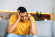 © Kateryna - Confused sad latino guy sitting on a sofa in the living room, dressed in casual yellow t-shirt, bewildered, talking on the phone, angry, annoyed, have unpleasant telephone conversation