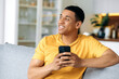 © Kateryna - Pensive happy young latino man in a yellow casual t-shirt, sitting at home on the couch, holding his smartphone in his hand, texting online, browsing social networks, looking away, smiling