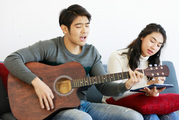 Portrait of cute smiling young Asian lover couple in white and gray long sleeve sweatshirt and blue jeans holding an acoustic guitar while using a digital tablet to search guitar notes on the internet