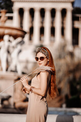 Portrait of young rich gorgeous redhead woman in sunglasses and summer dress standing sideways in ancient garden of old palace at fountain in Peterhof, Saint Petersburg looking at camera