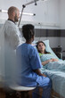© DC Studio - Medical team monitoring sick woman explaining sickness symptom examining medical recovery in hospital ward. Patient resting in bed while doctor writing treatment during rehabilitation appointment