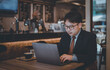 © APchanel - Asian businessman wearing suite and eyeglasses drink coffee , typing notebook for work at cafe restaurant.