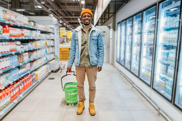 Wall Mural - Smiling African-American man with shopping basket stands between rack and refrigerators in modern supermarket department