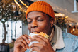 Pleasured African-America guy in orange knitted hat eats delicious fresh hamburger in contemporary decorated cafe close view