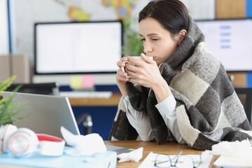 Sick young woman sits in warm blanket with cup at workplace