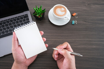 Wall Mural - Businessman hand holding blank note pad and pen on modern table desk background, Writing or recording with blank white notebook