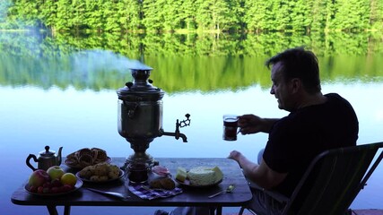 Wall Mural - Man drinking tea by the lake. Vintage metal tea samovar with white smoke and food on the table near the calm water lake in green forest at morning, Ukraine