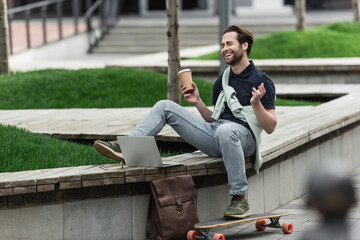 cheerful man in polo shirt and sweatshirt holding paper cup and laughing while sitting near longboard and gadgets.