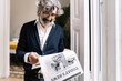 © COROIMAGE/Westend61 - Senior man with in-ear headphones reading newspaper at home