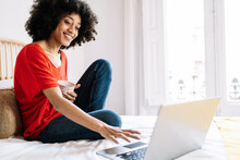 Young Woman With Mug Using Laptop While Sitting On Bed At Home