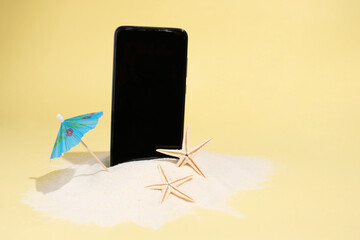 Smartphone in sand with umbrella and starfish on yellow background, summer concept, mock-up