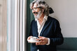 © COROIMAGE/Westend61 - Happy senior man with in-ear headphones holding coffee cup near window at home