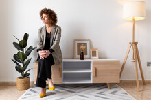 Mature Woman Looking Away While Sitting On Wooden Cabinet At Home