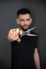 Young Male Barber Showing Scissor While Standing In Front Of Black Wall At Salon
