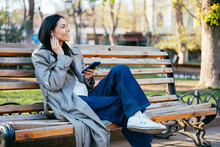 Smiling Woman Wearing Flare Pants Looking Away While Sitting On Bench In Park