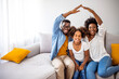 © Dragana Gordic - New building residential house purchase apartment concept. Stylish full family with kid sitting on sofa, mom and dad making roof figure with hands arms over head. Concept housing young family.