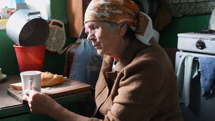 Wall Mural - Side view portrait of 80 years old woman having breakfast in rustic kitchen. Rural senior woman in traditional head scarf wrapped on her head. Life after retirement