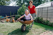 © Oxana Guryanova/Westend61 - Happy girl sitting in wheelbarrow while playing with grandmother at backyard