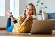 Contemplating Woman With Cryptocurrency Credit Card Looking Away While Lying Down In Front Of Laptop At Home