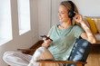 © steve brookland/Westend61 - Happy woman with headphones holding smart phone on armchair at home