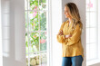 © steve brookland/Westend61 - Smiling woman leaning on wall looking through window in living room