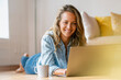 © steve brookland/Westend61 - Smiling woman lying on hardwood floor while using laptop at home