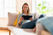 © steve brookland/Westend61 - Contented woman looking at digital tablet while sitting on sofa in living room
