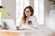 © steve brookland/Westend61 - Female professional with eyeglasses working on laptop while holding smart phone at desk