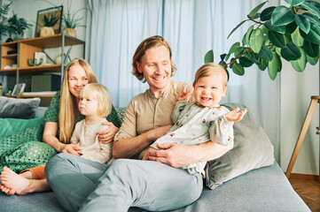 Happy young family of four relaxing on a couch, couple playing with baby girl and toddler boy